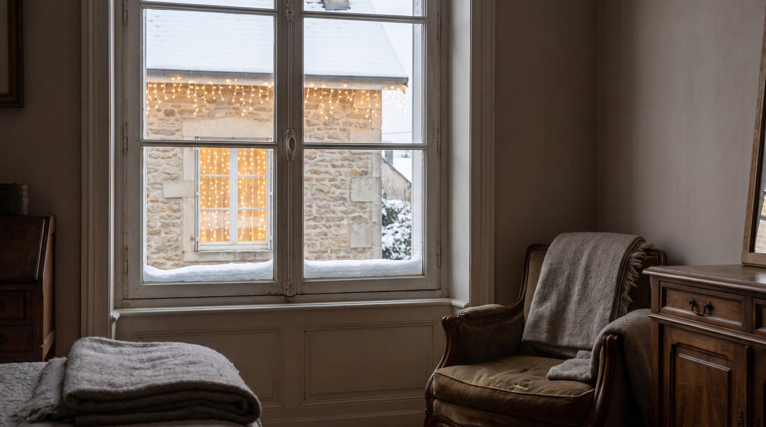 Façade de maison en pierre éclairée le soir par une guirlande lumineuse blanc chaud qui suit la ligne du toit, avec un rideau de lumière derrière une fenêtre et de la neige au sol.