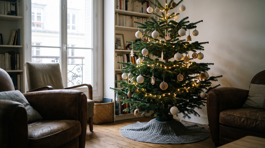 Un salon aux murs clairs en soirée, un sapin décoré de boules blanches mates et d'ornements en bois, une guirlande blanc chaud allumée près du tronc et un cache-pied gris au sol.